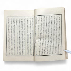 May include: An open book with Japanese text on aged, cream-colored pages. The text is in black ink, with vertical columns of characters. The book is slightly open, revealing two facing pages with detailed calligraphy and illustrations.