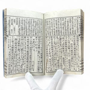 Puede incluir: Un libro abierto con páginas llenas de columnas verticales de texto japonés. El libro tiene una columna vertebral azul y está abierto por una mano enguantada. El papel está envejecido con un tono amarillento.