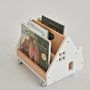 Könnte beinhalten: Ein weißes, hausförmiges Bücherregal aus Holz mit Innenpaneelen aus Naturholz und vier kleinen Lenkrollen. Das Seitenteil ist mit einem ausgeschnittenen Fenster- und Türdesign versehen. Mehrere Kinderbücher stehen aufrecht im Regal.