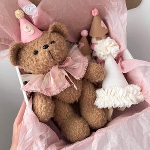 Pode incluir: Um urso de peluche castanho com um chapéu de festa cor-de-rosa com o número 9 e um colarinho de folhos cor-de-rosa, numa caixa com papel de seda cor-de-rosa, juntamente com dois chapéus de festa adicionais, um bege com o número 1 e um branco.