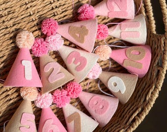 Puede incluir: Un conjunto de doce gorros de fiesta cónicos de fieltro en tonos rosa y beige. Cada gorro presenta un número de fieltro del uno al doce, un pequeño pompón en la parte superior y una fina cuerda elástica para sujetarlo bajo la barbilla.