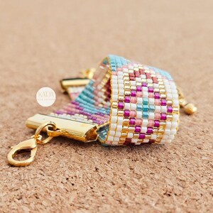 May include: A beaded bracelet with a gold clasp. The bracelet features a geometric pattern in shades of white, pink, purple, and turquoise. The clasp and connecting hardware are gold-toned. The brand name "KATJA" is visible on a small white circle.