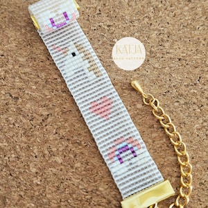 May include: A beaded bracelet with a white background and a pixelated design of a unicorn, hearts, and geometric shapes. The bracelet has gold-colored clasps and chain. The text "KATJA BEAD PATTERN" is in a circle in the background.