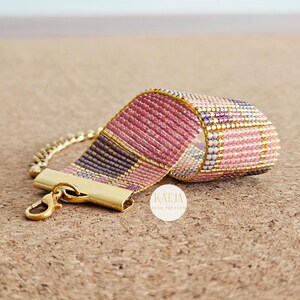 May include: A woven bead bracelet featuring a plaid pattern in shades of pink, purple, and gold. The bracelet includes a gold-tone metal clasp and a short chain extension. A small white circular logo with the text KATJA BEAD PATTERN is visible.