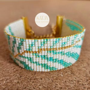 Peut inclure: Un bracelet de perles avec un motif géométrique blanc, turquoise et doré. Le bracelet a un fermoir doré et une rangée de perles dorées. Le texte "KATJA BEAD PATTERN" est sur un cercle blanc en arrière-plan.