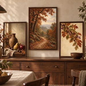 May include: A set of three framed wall art prints featuring autumn themes. The left print shows a still life with a ceramic jug, pear, and apple. The centre print depicts a forest path with orange trees. The right print displays oak leaves and acorns.