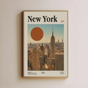 May include: A framed poster featuring a New York City skyline with the text "New York" at the top. The image includes the Empire State Building and a large orange circle. The poster has a beige background and a brown frame.