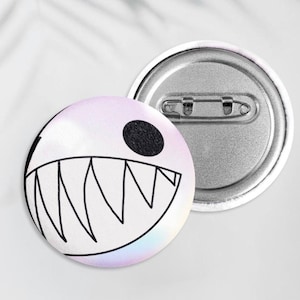 Puede incluir: Dos insignias redondas. Una insignia presenta un diseño de dibujos animados de una boca con dientes afilados y un ojo de punto negro sobre un fondo blanco con un sutil brillo de arcoíris. La otra insignia es plateada con un alfiler de seguridad.