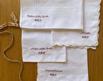 May include: Four white fabric handkerchiefs with scalloped or straight edges, featuring brown embroidered text. The text reads Father of the Bride, Mum, Mother of the Bride, and Maid of Honour, each followed by the date 02.02.27.