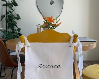 May include: A white rectangular fabric sign with frayed edges and the word Reserved printed in black script. The sign features metal grommets at the top corners and is tied to the back of a yellow wooden chair with white fabric strips.