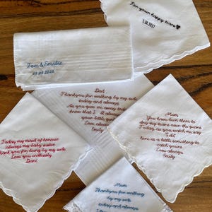 May include: A collection of white fabric handkerchiefs featuring various embroidered messages in blue, red, and brown thread. Some handkerchiefs have scalloped edges, while one features a delicate white lace trim. Each piece includes personalised text.