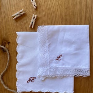 Könnte beinhalten: Zwei weiße quadratische Taschentücher mit gewelltem Rand und Spitzenbesatz. Ein Taschentuch zeigt das braun gestickte Monogramm JE, während das andere den braun gestickten Buchstaben L aufweist. Drei kleine hölzerne Wäscheklammern liegen daneben.