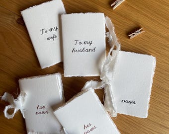May include: Five small, off-white handmade paper booklets with deckled edges and frayed ribbon ties. The covers feature black embroidered text reading To my wife, To my husband, her vows, his vows, and vows. Four small wooden clothespins are included.