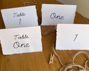 May include: Four white rectangular folded cards with deckled edges featuring black handwritten text. The cards display the phrases Table 1, One, Table One, and the number 1. A length of brown jute twine and two small wooden clothespins are included.