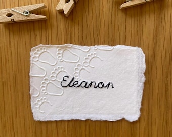 May include: A rectangular piece of white handmade paper with deckled edges, featuring an embossed pattern of baby footprints and the name Eleanon embroidered in black thread, accompanied by three small wooden clothespins.