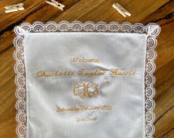May include: A square white fabric handkerchief with a decorative lace border. The center features gold embroidery reading Welcome, Charlotte Taylor Harris, the initials CTH, Saturday 8th June 2028, and God Bless. Four small wooden clothespins are nearby.