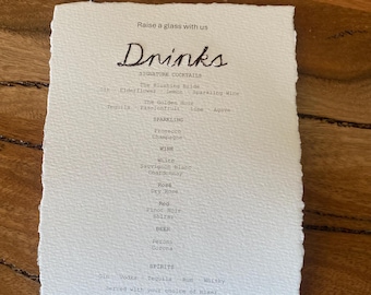 May include: A white, rectangular paper menu with deckled edges featuring black printed text. The header reads "Raise a glass with us" above the word "Drinks" in a script font, followed by lists of signature cocktails, sparkling, wine, beer, and spirits.