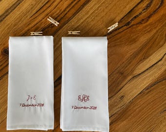 May include: Two white rectangular fabric napkins featuring red embroidery. The left napkin displays the text J+E and 7 December 2028. The right napkin features a monogram with the letters SHE and the text 7 December 2028. Four small wooden clips are included.