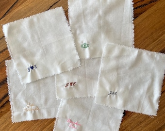 May include: A collection of six square white fabric napkins with frayed edges. Each napkin features a unique embroidered monogram or name in various thread colors, including blue, green, dark red, black, light pink, and cream, positioned in the corner.