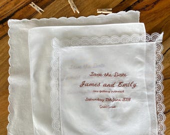 May include: Three white square handkerchiefs with scalloped or lace edges. The top handkerchief features embroidered text in a reddish-brown thread that reads: Save the Date, James and Emily are getting married, Saturday 12th June, 2028, Gold Coast.