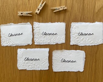 May include: Five rectangular white paper cards with deckled edges, each featuring the name Eleanon embroidered in black thread. The cards display various embossed patterns including floral designs and baby footprints, accompanied by three wooden clothespins.