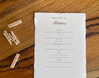 May include: A rectangular white menu card with deckled edges featuring the text Dine with us Menu followed by a list of food items including seared scallops, beef tenderloin, and panna cotta, accompanied by three light brown wooden clothespins.