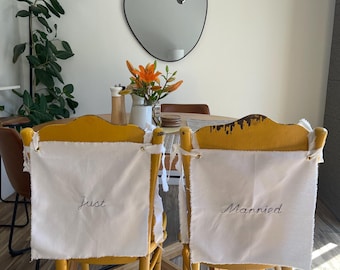 May include: Two yellow wooden chairs with distressed paint, each featuring a white fabric cover tied to the backrest. The left cover is embroidered with the word "Just" and the right cover is embroidered with the word "Married" in black script.