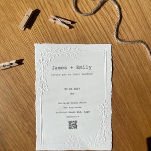 May include: A rectangular white wedding invitation card with deckled edges, featuring embossed floral corner designs and black typewriter-style text reading James + Emily, Invite you to their wedding, 07.02.2027, 4pm, Burleigh Heads Beach, Australia.