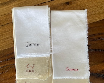May include: Two rectangular strips of off-white fabric with frayed edges featuring embroidered text. One strip displays the name James, E+J, and 13.02.27. The other strip displays the name Emily + James, 13.02.2027, the name Emma, and the word Bride.