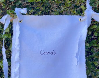 May include: A square white fabric sign with frayed edges featuring the word "Cards" embroidered in black cursive script. The sign includes two gold-toned metal grommets at the top corners, each threaded with long, white, raw-edged fabric ties.