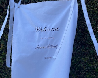 May include: A white fabric banner with frayed edges and long fabric ties at the top corners. The text printed in black ink reads: Welcome to our wedding, James & Emily, 18.10.2027. The banner is rectangular and hangs vertically.
