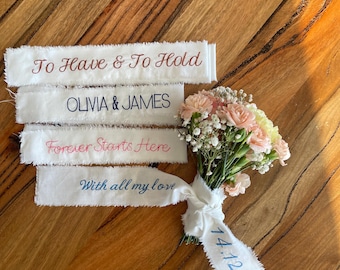 May include: A small floral bouquet of light pink carnations and white baby's breath tied with white fabric ribbons. The ribbons feature embroidered text: To Have & To Hold, OLIVIA & JAMES, Forever Starts Here, With all my love, and 14.12.2027.