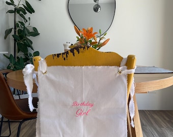 May include: A white fabric chair cover with frayed edges and gold-toned metal grommets at the top corners. The text "Birthday Girl" is embroidered in pink script on the center of the fabric. The cover is tied to a yellow wooden chair.