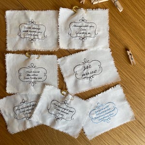 Puede incluir: Seis parches de tela rectangulares de color blanco roto con bordes deshilachados, cada uno con texto bordado en negro o azul dentro de un marco decorativo. Los parches incluyen imperdibles dorados y tres pinzas de madera pequeñas.