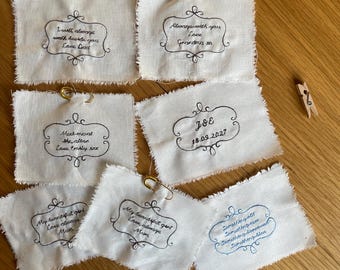 May include: Six rectangular off-white fabric patches with frayed edges, each featuring black or blue embroidered text inside a decorative frame. The patches include gold safety pins and are arranged with three small wooden clothespins nearby.