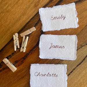 Puede incluir: Tres tarjetas rectangulares de papel blanco con bordes irregulares y deshilachados, cada una con un nombre bordado en hilo marrón. Los nombres son Emily, James y Charlotte. Tres pequeñas pinzas de madera están situadas a la izquierda.