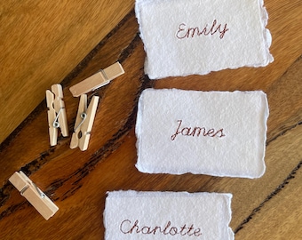 May include: Three rectangular white paper cards with rough, deckled edges, each featuring a name embroidered in brown thread. The names are Emily, James, and Charlotte. Three small wooden clothespins are positioned to the left of the cards.