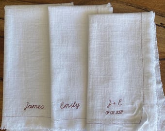 May include: Three white linen napkins with frayed edges and brown stitched borders. Two napkins are embroidered with the names James and Emily, while the third features the initials J + E and the date 07 02 2027 in brown thread.