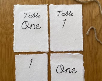 May include: Four rectangular white paper cards with deckled edges featuring black embroidered text. The cards display the words Table One, Table 1, 1, and One. Three small wooden clothespins and a length of jute twine are positioned above.
