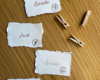May include: Four rectangular white paper cards with deckled edges featuring embroidered names and dietary icons. The cards display the names Bride, Jack, Bride, and Ione, accompanied by symbols for no shellfish, no gluten, vegan, and vegetarian diets, alongside two wooden clips.