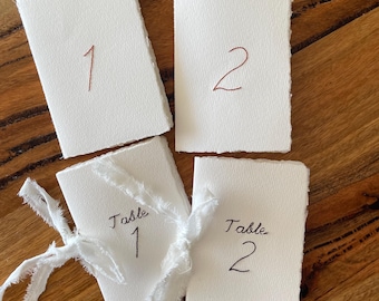 May include: Four white rectangular folded cards with deckled edges. Two cards feature the embroidered numbers 1 and 2 in brown thread. Two cards feature the embroidered text Table 1 and Table 2 in black thread, with one tied with white ribbon.