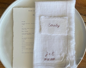 May include: A white linen napkin with frayed edges featuring brown embroidered text that reads J + E and 07 02 2027, topped with a small white card with deckled edges displaying the name Emily in brown script, resting on a menu and a white plate.