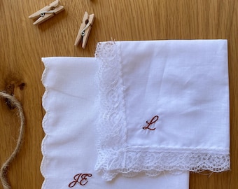 May include: Two white cotton handkerchiefs with scalloped edges and delicate lace trim. One handkerchief features the embroidered brown monogram JE, while the other displays the embroidered brown letter L. Three small wooden clothespins are included.
