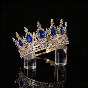 Könnte beinhalten: Eine goldene Krone mit einer Reihe von tropfenförmigen blauen Kristallen und klaren weißen Strasssteinen, die in einem dekorativen Filigranmuster angeordnet sind. Die Krone hat eine kreisförmige Basis und mehrere Spitzen mit kleinen Kristallen.