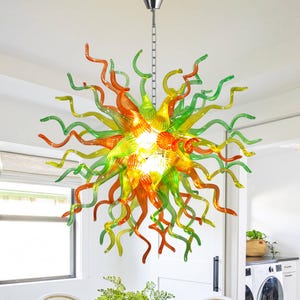 May include: A colorful, artistic chandelier with a sunburst design. The fixture features a central cluster of lights surrounded by swirling, elongated glass tendrils in shades of orange, yellow, and green. It hangs from a silver chain.