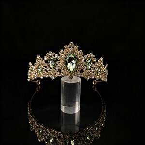 Peut inclure: Un diadème doré orné d'un cristal central vert clair en forme de poire, entouré de cristaux verts assortis plus petits et de nombreux strass transparents sertis dans une monture en filigrane métallique au design pointu.