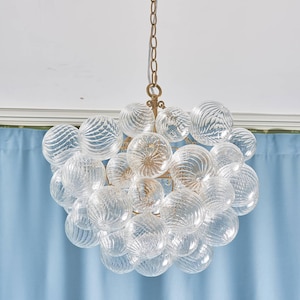 Peut inclure: Un lustre en verre transparent avec de multiples ampoules sphériques. Les ampoules ont un motif texturé et tourbillonnant et sont disposées en grappe. Le lustre est suspendu à une chaîne et un luminaire de couleur dorée.