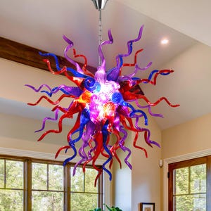 Peut inclure: Un lustre artistique et coloré avec un design en forme d'éclat d'étoile. Le luminaire présente des éléments en verre torsadés et allongés dans des tons rouges, bleus et violets. Le luminaire est suspendu au plafond par une chaîne argentée.
