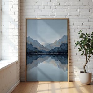 May include: A framed landscape painting shows a mountain range reflected in water. The artwork uses blue and grey tones, with a pale sky. The frame is light wood, and the painting is displayed against a white brick wall. The painting is approximately 100cm x 70cm.