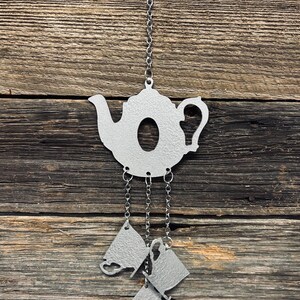 May include: A silver-colored metal teapot-shaped wind chime with three dangling teacup charms. The teapot has a handle and spout, and hangs from a chain and hook. The background is a rustic wooden surface.